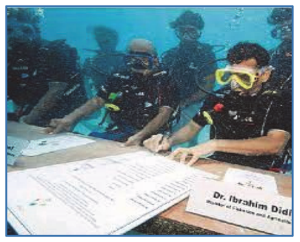 Fotografia ilustrująca tekst „Underwater Cabinet Meeting”: ministrowie w strojach do nurkowania siedzą przy stole pod wodą.